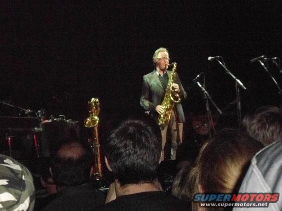 Man in a suit playing a saxophone on a dimly lit stage with microphones