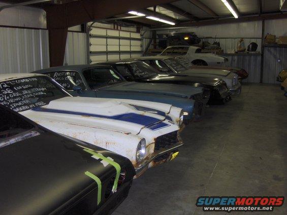 Several old cars lined up in a garage. One has white paint with blue stripes and writing on the hood