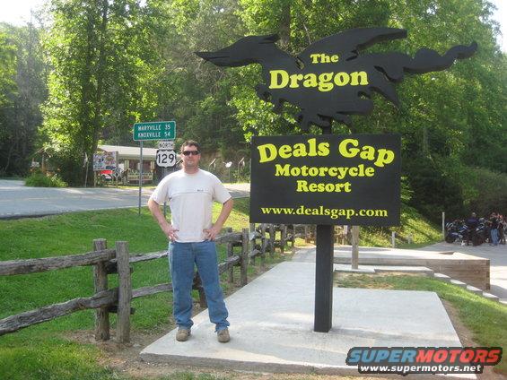 Man in t-shirt and jeans stands by the Deals Gap Motorcycle Resort sign. Road signs for Maryville and Knoxville are visible