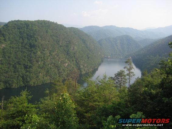 Scenic mountain lake with a dam and trees in the foreground. Watermark reads SUPERMOTORS.NET