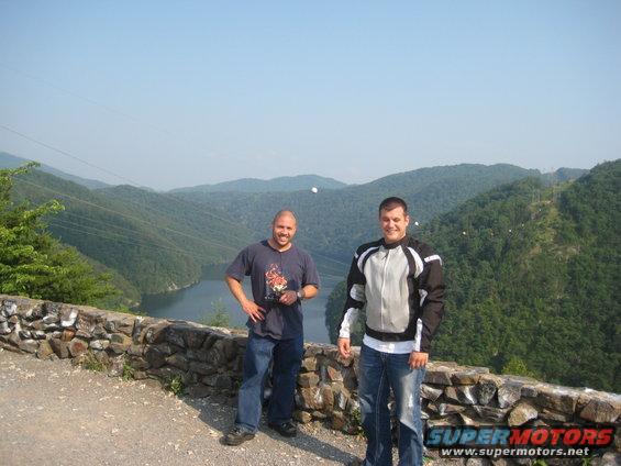 Two men stand on a stone wall overlooking a wide, tree-covered valley with a river below. One man wears a t-shirt and jeans, the other a motorcycle jacket and jeans