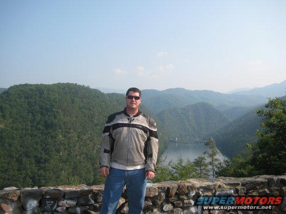 Man in gray and black motorcycle jacket and blue jeans stands on stone wall overlooking a lake and mountains