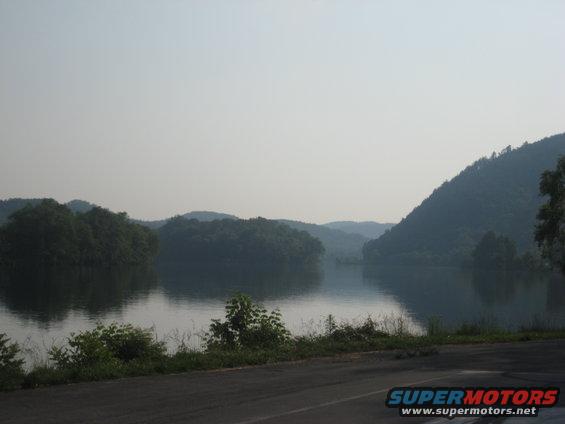 Water body with tree-covered hills under a hazy sky, asphalt road in foreground