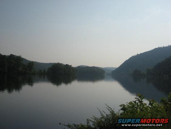 Calm lake reflects tree-covered hills under a hazy sky
