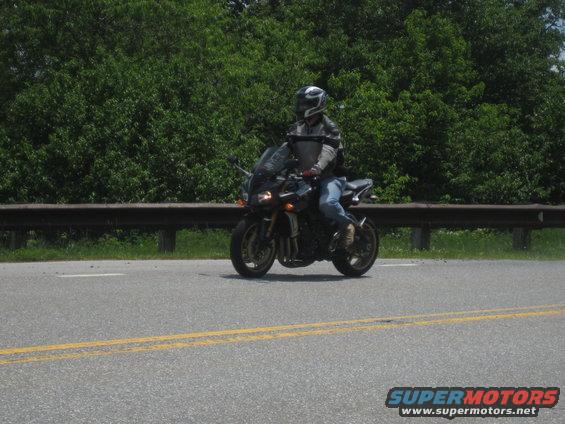 Motorcyclist wearing a helmet and leather jacket rides a dark sportbike on a paved road with trees in the background