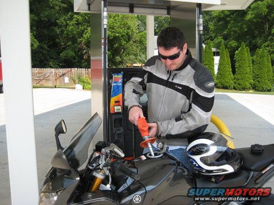 Black motorcycle at gas pump with man fueling. Helmet on seat