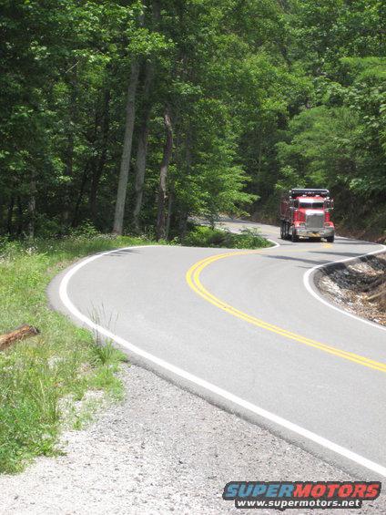 Red dump truck driving on a two-lane road with yellow dividing lines, surrounded by trees