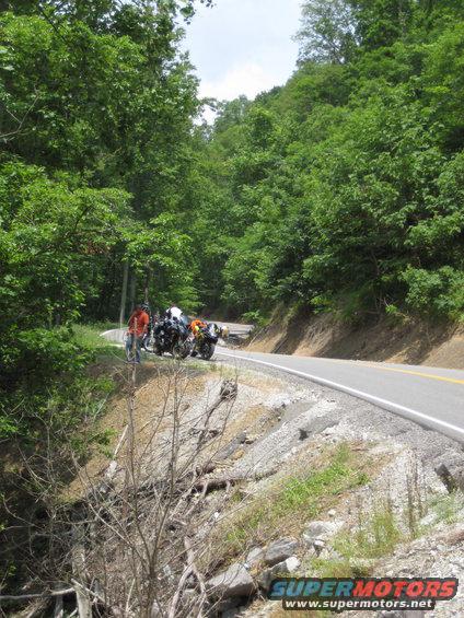 Two motorcycles parked on the side of a winding mountain road with trees on either side