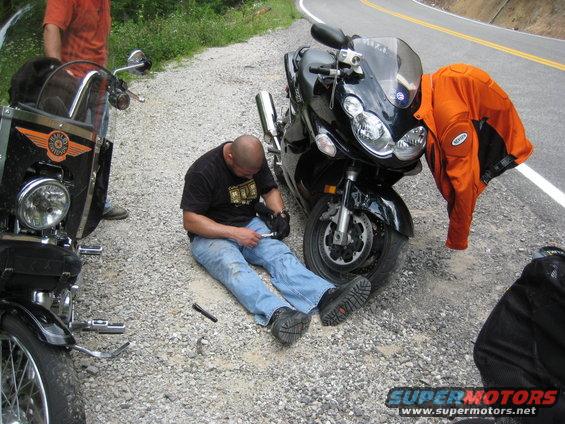 Guy fixing a motorcycle on the side of the road next to another bike. Orange jacket on the bike