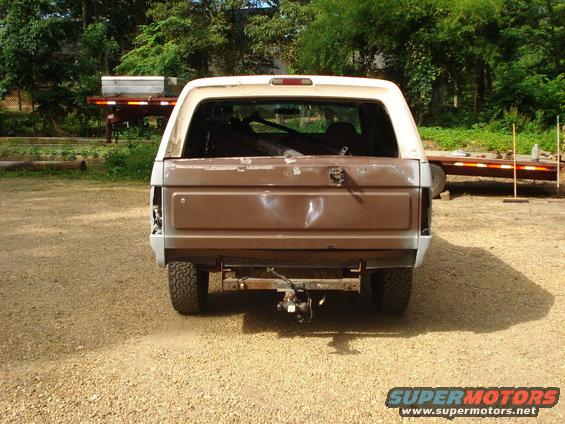 Brown and white Ford Bronco, rear view, dented tailgate, missing taillight, parked on gravel