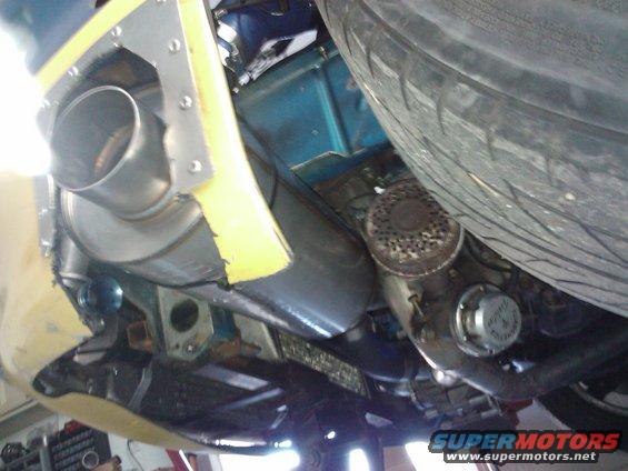 Underside of a car showing exhaust pipe, part of the drivetrain, and a tire. Some yellow tape is visible on the body panel