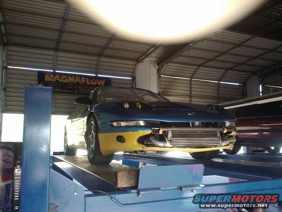 Blue and yellow car on a lift, front view, with exposed intercooler and aftermarket headlights