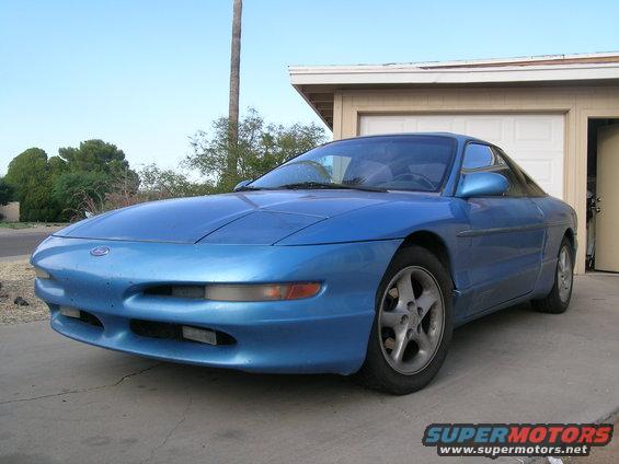 Blue coupe parked in front of a garage, front 3/4 view. The car has a dented front bumper and a visible 'SUPERMOTORS.NET' watermark