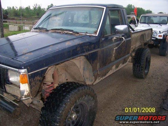 Muddy dark blue Jeep pickup truck parked next to a white Jeep Wrangler. Both have large off-road tires