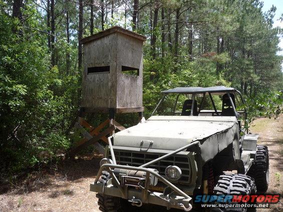 Olive drab off-road vehicle with roll cage, winch, and large tires, parked next to a wooden deer stand