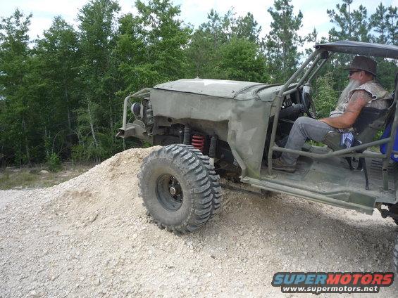 Custom off-road buggy with large tires, driven by a man wearing a hat and vest, on a dirt mound