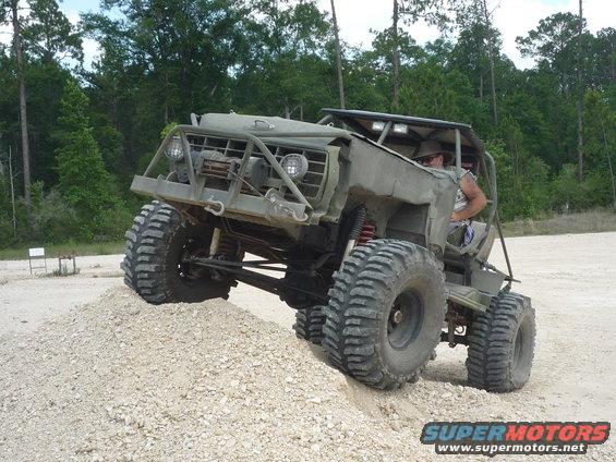 Olive drab off-road vehicle with large tires climbing a dirt mound, front grille and winch visible