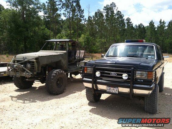 Front view of a black 1990 Ford Bronco with a brush guard and a modified off-road vehicle parked next to it