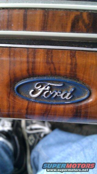 Ford emblem on wood grain dash trim. Jeans and sneakers visible below