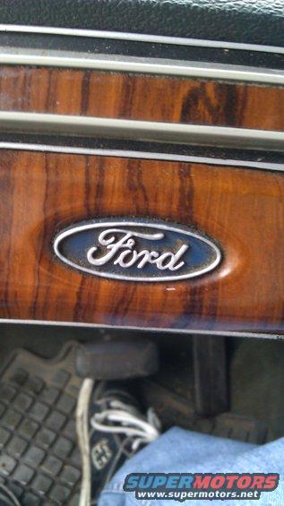 Ford emblem on wood-grain trim inside a vehicle, with a foot on the pedal