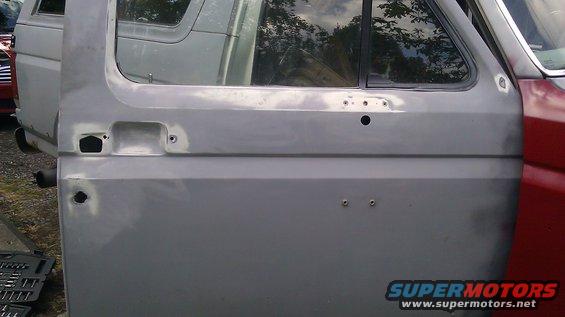 Gray primer door panel with holes for handle and mirror, red fender on right. Another vehicle visible behind