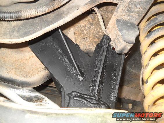 Close-up of a black, welded metal part attached to a vehicle's suspension system, near a yellow coil spring