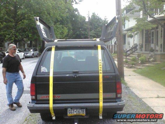 Rear view of a black GMC Jimmy with gullwing doors open, secured by yellow straps. A man stands to the left