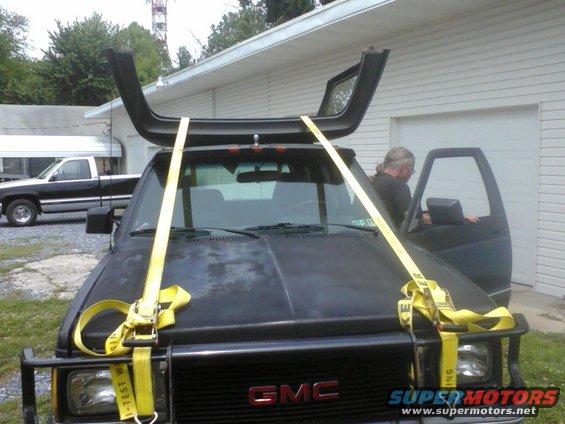 Black GMC truck with yellow straps securing a black roof rack part to its hood; a person is in the driver's seat