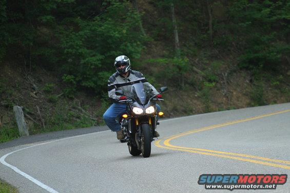 Guy on a dark sportbike riding on a curvy road with trees on the side. The bike has dual headlights