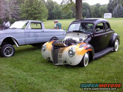 Hot rod coupe with flame paint on front fender, and a light blue pickup truck parked on grass