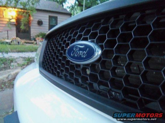 White car's front grille with a Ford logo. Honeycomb pattern. House in background