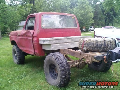 Red pickup truck chassis with a spare tire on the back. Another vehicle is partially visible to the right
