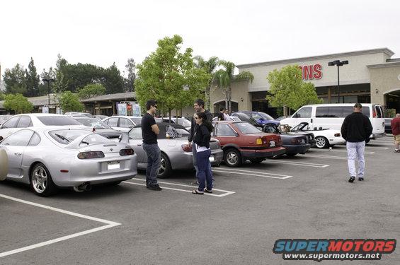 Parking lot scene with several cars including a silver sports car, a red sedan, and a white van. People are standing and walking
