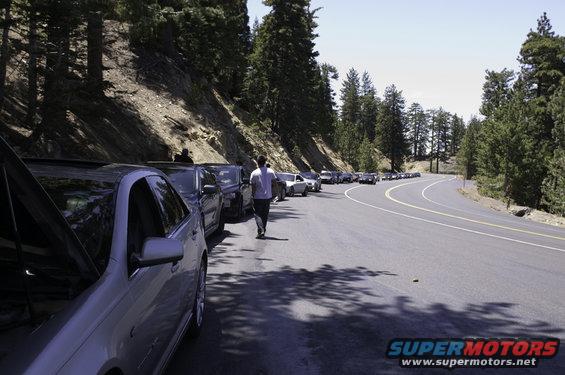 Cars parked along a winding mountain road with trees on the left and a clear sky