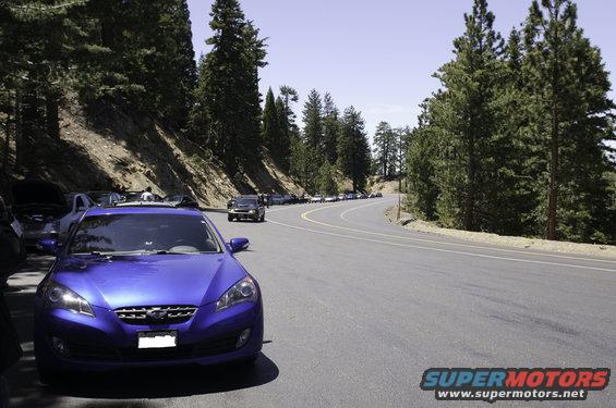Front view of a blue Hyundai Genesis sedan parked on the side of a winding mountain road with other cars in the background