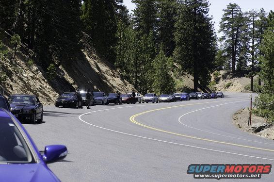 Blue car side mirror and hood in foreground, line of cars parked on winding mountain road with trees and rocky slope