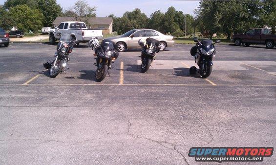 Four motorcycles parked in a lot with cars and trucks in the background