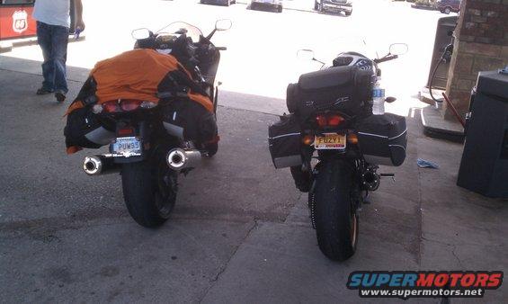 Two motorcycles parked side-by-side at a gas station, both loaded with saddlebags and top cases. One has an orange cover on the rear
