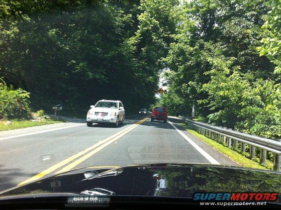 View from inside a car looking out at a two-lane road with a white SUV and a red car ahead, surrounded by trees
