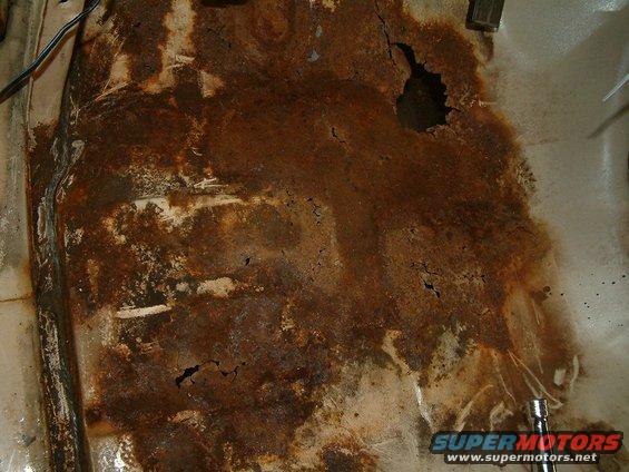 Rusted floor pan with holes, showing extensive corrosion and some white undercoating