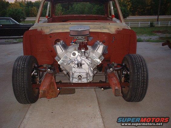 Front view of a vehicle chassis with a V8 engine installed, showing exposed red metal body panels and suspension components