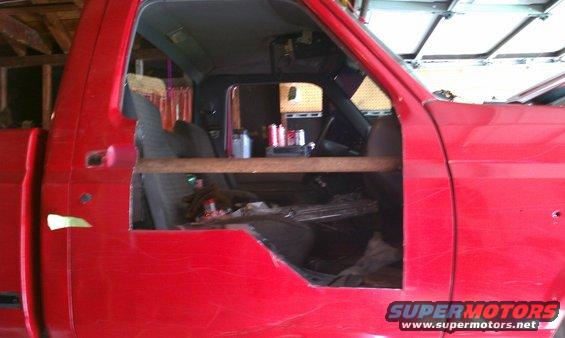 Red pickup truck cab with door removed, showing seats and dashboard. A wooden bar is across the opening