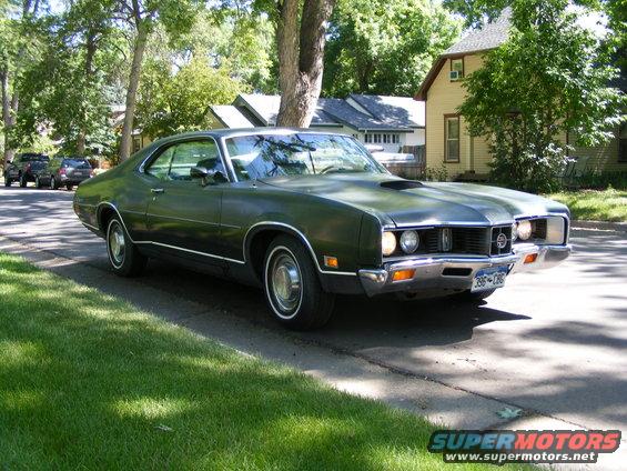 Dark green 1970 Mercury Cougar XR7 coupe parked curbside, front 3/4 view. Hood scoop, chrome bumper, whitewall tires
