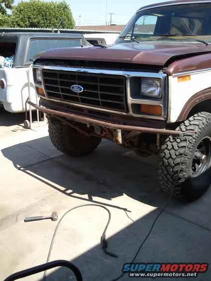 Front of a brown and white Ford F-Series truck with a custom bumper. Another vehicle is parked behind it
