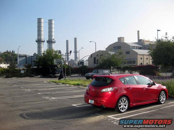 Red hatchback parked in a lot with a power plant in the background. Another car is parked nearby