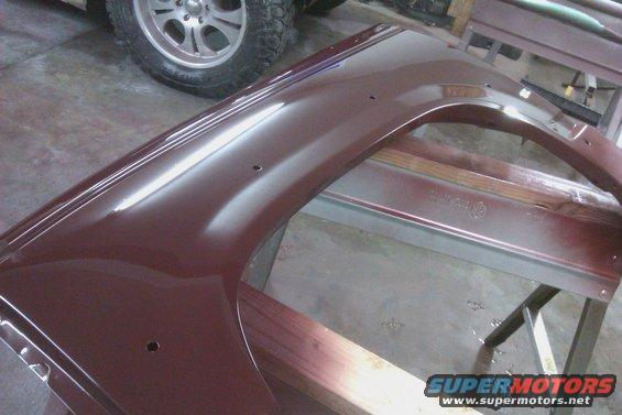 Maroon car fender with three mounting holes, resting on sawhorses. A tire and wheel are visible in the background