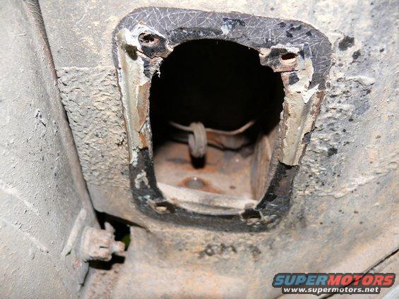 Underside of a vehicle showing a rusted, open access panel with a wire loop inside