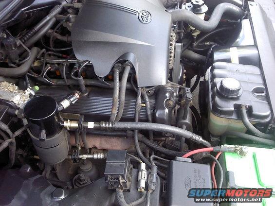 Engine bay with V8 emblem on cover. Shows battery, hoses, and wiring