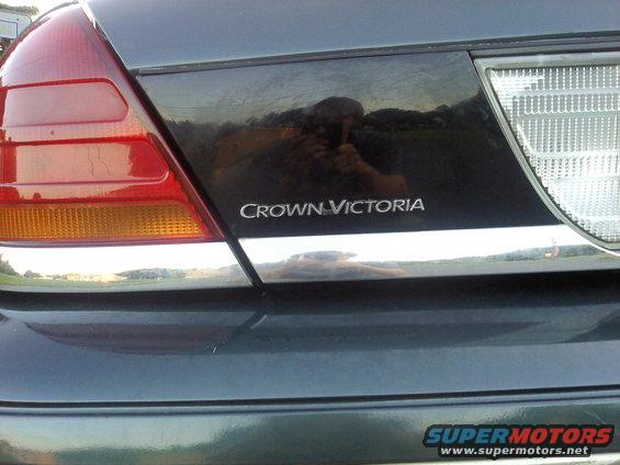 Dark-colored vehicle's rear with a chrome trim strip below the Crown Victoria emblem and a red taillight