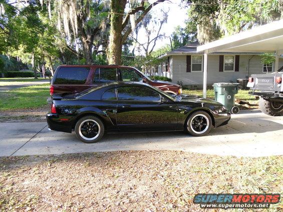 Black Ford Mustang coupe parked on driveway, side view. Another SUV and a Jeep are also visible